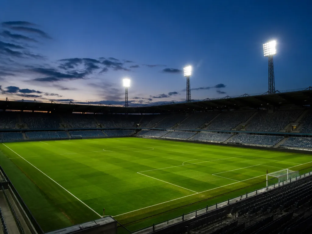 Tom fotbollsplan i skymning med tända strålkastare och tomma läktare i bakgrunden